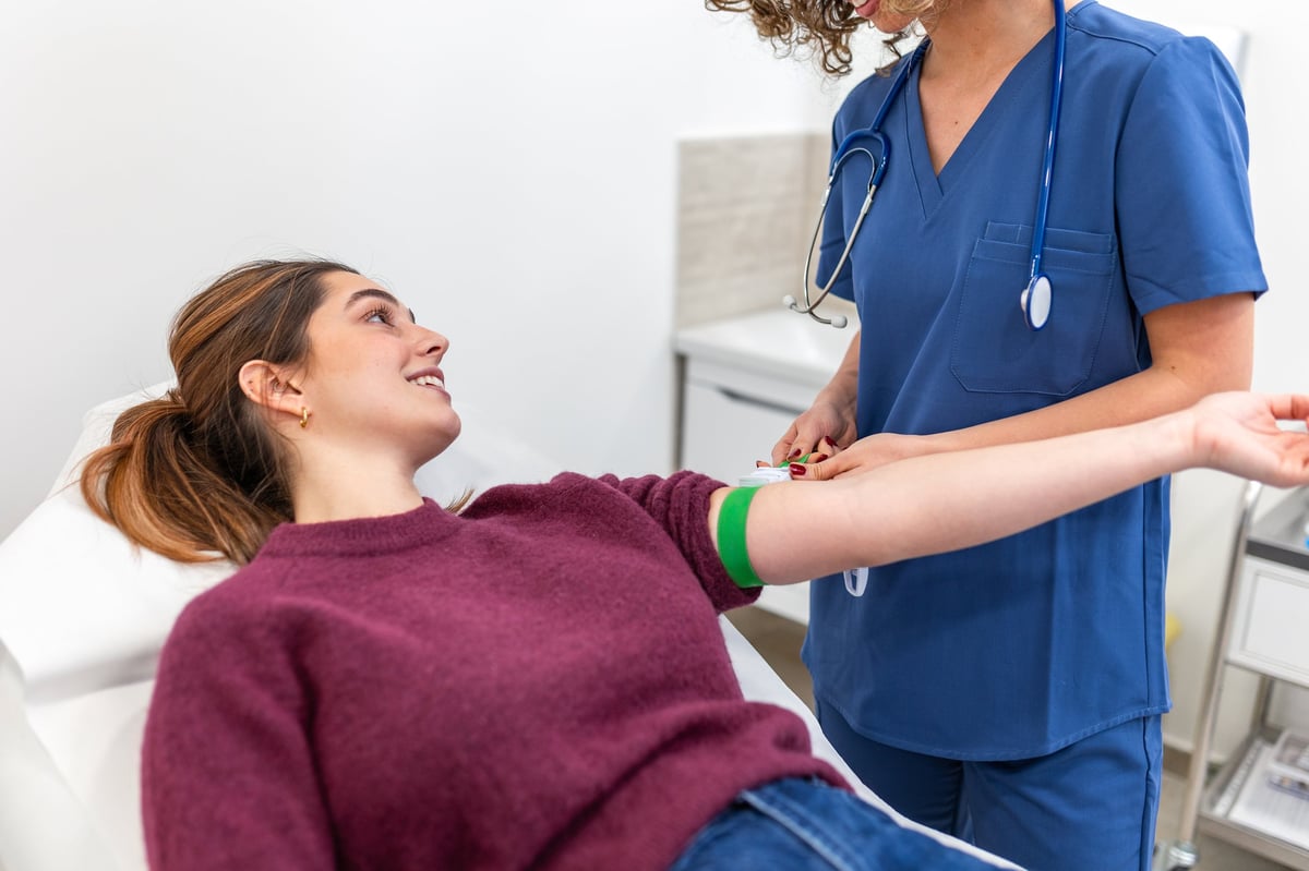 A phlebotomist in blue scrubs preparing a smiling patient's arm with a tourniquet for a blood draw